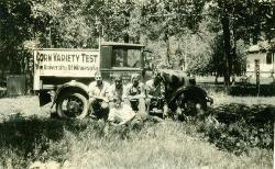 black and white photo of four people sitting next to a corn variety test truck
